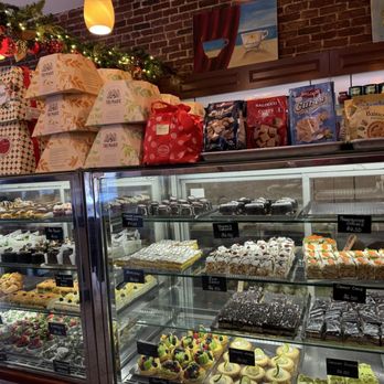Modern Pastry Shop - A bakery display case contains various cakes and pastries, with boxes and gift bags on top, and a brick wall with artwork in the background. - Bakeries Near Me - Boston, Massachusetts