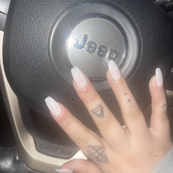 Arts Nails - Length and shape - A hand with long, white acrylic nails and tattoos on fingers rests on a Jeep steering wheel inside a vehicle. - Nail Salons Near Me - Lewiston, Maine