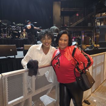 Rams Head Live - The impersonator and I - Two women are positioned behind a metal barrier in front of a stage with musical instruments and sound equipment. One woman wears a white lace shirt and has curly hair, while the other wears a red jacket and black pants. The background shows stage crew members preparing for a performance in a concert venue with multiple levels and stairs visible on the right side. - Music Venues Near Me - Baltimore, Maryland