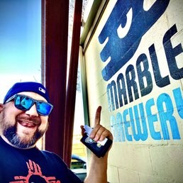 A man with a beard, wearing reflective sunglasses and a baseball cap, is smiling broadly and pointing at a sign that reads 'MARBLE BREWERY.' The sign is mounted on a wall outside the brewery, which features a wooden structure and a clear sky in the background. The man appears to be enjoying his visit to the brewery.