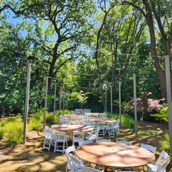 Sonoma Party Rentals - Outdoor event setup featuring round tables, white folding chairs, and bistro lighting nestled among the trees.