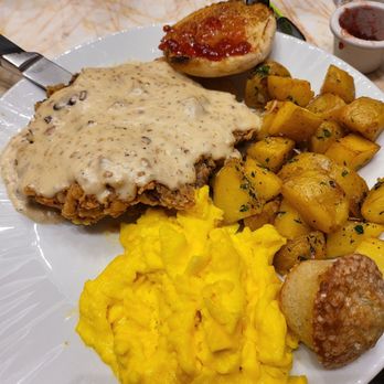 Biscotti's - Chicken fried steak can eggs home style potatoes - A breakfast plate includes chicken fried steak topped with creamy gravy, scrambled eggs, roasted potatoes garnished with herbs, and a baked potato with ketchup. The meal is served on a white ceramic plate, with a table setting that includes glasses, utensils, and condiments in the background. - Cafes Near Me - Reno, Nevada