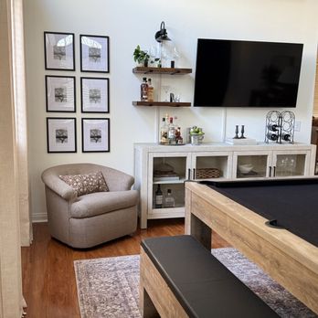 John Belanger Construction - A living space features a beige armchair with a decorative pillow, wall art of trees, and a white bar cabinet with glassware and bottles, adjacent to a pool table. - Handyman Near Me - Corona, California
