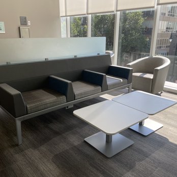 Rush South Loop - The image shows a waiting area with a black sofa, a beige armchair, and two white tables positioned in front of large windows. The space is illuminated by natural sunlight, and the windows reveal an urban view with trees and buildings outside. The area has a neutral color palette and a clean, organized appearance. - Doctors Near Me - Chicago, Illinois
