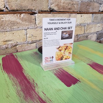Chit Chaat - A clear acrylic menu stand on a painted table features a naan and chai set at Chit Chaat restaurant. The table has a green base with red and yellow brush strokes. The background consists of a beige brick wall, creating a rustic ambiance. The menu includes images of naan bread and a cup of chai, with descriptions and pricing. - Indian Near Me - Racine, Wisconsin