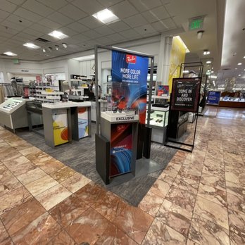 Macy's - A retail kiosk at Macy's in Albuquerque, NM, features sunglasses displays and promotional signs, with store fixtures and clothing racks visible in the background. - Department Stores Near Me - Albuquerque, New Mexico