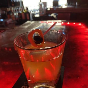 The Overpour - This is an Old fashion post prohibition, awesome! - A cocktail featuring a lemon slice and an olive on a pick is set on a bar counter illuminated by red lighting. The drink is in a short, clear glass, and the background shows a bar with bottles and glasses, slightly out of focus. The setting suggests a bar environment with ambient lighting. - Cigar Bars Near Me - El Paso, Texas