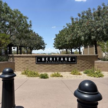 Big Chicken - Heritage Marketplace's sign is mounted on a brick wall, with trees and a clear blue sky behind it, and black bollards in the foreground. - Burgers Near Me - Gilbert, Arizona