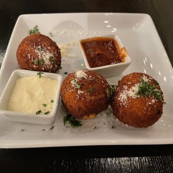 Debajo Tapas ABQ - Croquetas de Jamón y Pollo - Three croquetas are arranged on a white square plate, each topped with grated cheese and herbs. Two small square dishes contain dipping sauces, one creamy and the other dark and thick. The plate is placed on a dark wooden table, and the presentation suggests a Spanish-inspired tapas dish. - Spanish Near Me - Albuquerque, New Mexico