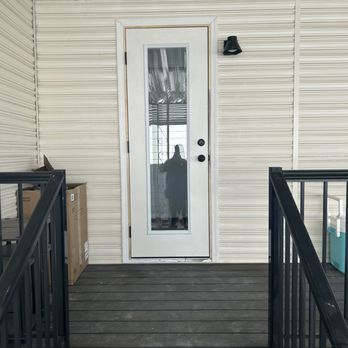 Response Crew - The image shows a white exterior door with a vertical glass panel, set in a beige siding wall. The door has a black handle and lock, and a black outdoor light fixture is mounted above it. The small porch area has dark wooden flooring and black metal railings on both sides. There are cardboard boxes on the left side of the porch, and a glimpse of a window with curtains can be seen through the glass panel in the door. - Handyman Near Me - Scottsdale, Arizona