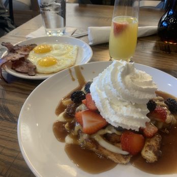 Another Broken Egg Cafe - French Toast Cinnamon roll covered in berries, cream cheese, syrup, and whip cream. With a side of bacon, sunny side up eggs, and a mimosa. - Breakfast & Brunch Near Me - Flowood, Mississippi