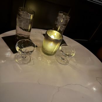 Rose Lounge - Adalina - Complimentary champagne - A round table with a white marble surface features two tall glasses of water, a lit candle in a textured metallic holder, and two empty cocktail glasses with stems. The setting appears dimly lit, with a dark background and soft lighting creating a cozy atmosphere. - Cocktail Bars Near Me - Chicago, Illinois