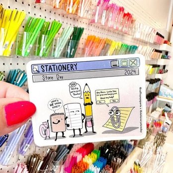 Calliope Paperie - The Founder of the Annual Stationery Store Day in August - Worldwide - A cartoon-themed stationery card featuring animated characters is held in front of a display of various colorful pens, markers, and stationery supplies at Calliope Paperie. The background shows organized shelves with pens in multiple colors, including green, pink, orange, and purple, arranged in containers. The card has a humorous illustration with speech bubbles and a title indicating Store Day 2024. - Gift Shops Near Me - Hudson, Massachusetts