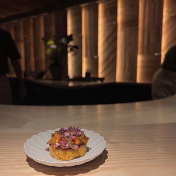 Ikigai - A small, round food item with a golden-brown base is topped with colorful ingredients, including purple and orange elements, and garnished with small purple flowers. It is served on a white scalloped plate placed on a light wooden counter. The background features a dimly lit interior with wooden paneling and a potted plant, with a person partially visible on the right side. - Japanese Near Me - Brooklyn, New York