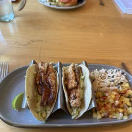 Fish Tacos - Two fish tacos with grilled fish, shredded lettuce, and lime are on a metal tray, accompanied by side salads with diced vegetables and pasta.