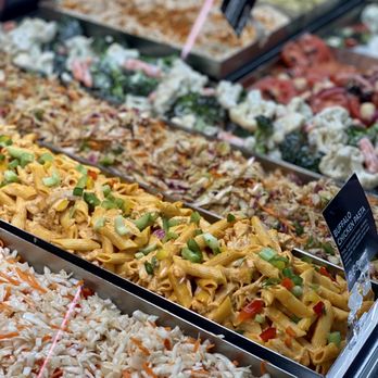 Hy-Vee - a display of various types of food - Multiple trays of prepared salads and pasta dishes are displayed in a grocery store. The dishes include pasta with chicken and vegetables, coleslaw, and other mixed salads. The trays are organized in a refrigerated display case, with small black signs indicating the names and prices of the items. The image captures the colorful and diverse selection of ready-to-eat food options available for customers. - Grocery Near Me - Lenexa, Kansas