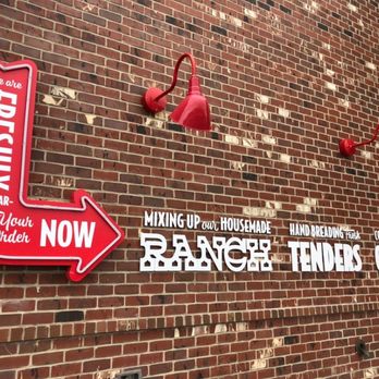Slim Chickens - Signage - The image shows a red neon sign shaped like an arrow with the words "We are FRESHLY PREPARING Your Order NOW" in white lettering, mounted on a brick wall. Two red wall-mounted lamps are positioned above the sign, and additional white text on the wall indicates steps for ordering, such as mixing, hand-breading, and cooking tenderized meat. - Fast Food Near Me - Avon, Ohio