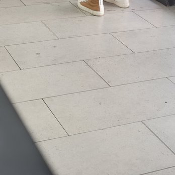 Hi BoBa - Dirty floor - A photograph of a tiled floor featuring rectangular, light-colored tiles with visible grout lines. The tiles have some dirt, small marks, and imperfections. At the top of the image, part of a person's legs and shoes are visible, with the shoes being white and brown sneakers. The setting appears to be indoors, possibly in a public or commercial space. - Bubble Tea Near Me - Albuquerque, New Mexico