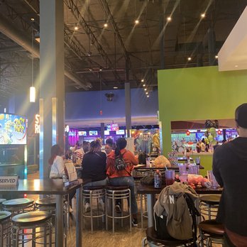 Main Event Gilbert - Visitors are seated at a bar counter in an arcade with gaming machines and colorful lighting in the background. - Bowling Near Me - Gilbert, Arizona