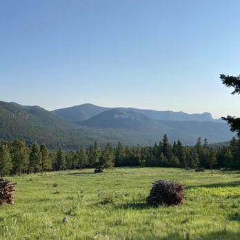 Philmont Scout Ranch - Meadow at crater lake - A wide view of a grassy meadow with stacks of firewood, surrounded by trees, with mountain ranges in the background and a clear sky. - Hiking Near Me - Cimarron, New Mexico