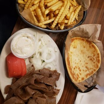 Dengeos - The image shows a meal consisting of a plate with sliced beef, sliced onions, and condiments including ketchup and a creamy sauce. There is a basket of crinkle-cut French fries and a separate basket with a slice of flatbread. The food is arranged on a wooden table, with utensils and napkins nearby. - Greek Near Me - Buffalo Grove, Illinois