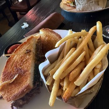 J Murphys on Main - Short rib, grilled cheese sandwich, french fries - A plate holds a grilled cheese sandwich with toasted bread and melted cheese, accompanied by a portion of French fries in a paper-lined basket. A small container of ketchup is placed on the side. The setting appears to be a casual dining environment with wooden tables and chairs, and another sandwich is visible in the background. - American Near Me - Cold Spring, New York