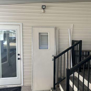 Response Crew - The photo depicts an exterior wall with a white door featuring a small rectangular window. To the left, there is a sliding glass door with a white frame. To the right, a staircase with black metal railings and wooden steps leads upward. The wall behind the door and stairs is made of beige, horizontally ridged siding. A small security camera or light fixture is mounted above the door. The ground is concrete, and part of a black outdoor chair is visible in the lower left corner. - Handyman Near Me - Scottsdale, Arizona