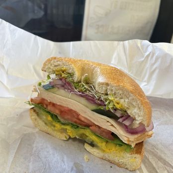 Bagel Street Cafe - Turkey bagel sandwich - A partially eaten bagel sandwich with turkey, tomato, cucumber, lettuce, sprouts, and mustard, placed on white paper. - Bagels Near Me - Berkeley, California