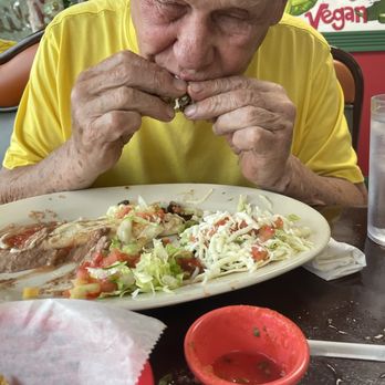 Cervantinos - Bobby absolutely loving his tacos - An elderly man in a yellow shirt is sitting at a table, eating a taco with his hands. There is a plate of food, a small bowl of sauce, and a glass of water on the table. - Mexican Near Me - Palos Heights, Illinois