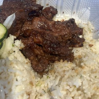 Chee-Bog - Tapsilog - The image depicts a close-up of a meal in a white plastic container, featuring cooked beef with a dark, glossy sauce, accompanied by white rice and slices of cucumber. The beef appears tender and well-seasoned, with some visible fat and caramelization. The rice is fluffy with individual grains visible, and a few cucumber slices are partially visible on the side. The container has a textured interior surface, and some sauce or food residue is present on the sides. - Filipino Near Me - Cohoes, New York