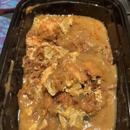 Shrimp Egg Foo Young. - A black plastic container contains shrimp egg foo young, featuring a mixture of eggs, shrimp, and vegetables covered in a thick gravy. The dish appears partially eaten, with some gravy pooled at the bottom. The background includes a colorful fabric surface, indicating it was served as takeout food from a Chinese restaurant.