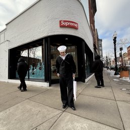 Supreme Chicago - A person dressed in a sailor outfit, including a white sailor hat and dark clothing, stands on the sidewalk in front of a Supreme store. The store has a white brick exterior with a red Supreme sign. Two other individuals are seen walking or standing nearby, and the city street features lampposts, buildings, and some snow on the ground. The sky is partly cloudy.