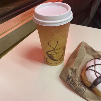 King Donuts - A paper coffee cup with a lid and a glazed donut with chocolate drizzle are placed on a table near a window. - Donuts Near Me - Newington, Connecticut