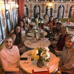 Celebrating the soon-to-be-bride.... thanks to the staff at Odelia's - A group of women is seated around a round dining table in a restaurant with elaborate wallpaper depicting classical sculptures and architectural elements. The table is decorated with a floral centerpiece, and the women are smiling and engaged in conversation. The setting includes warm lighting, curtains, and a sophisticated interior design.