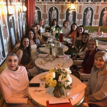 Odelia - Celebrating the soon-to-be-bride.... thanks to the staff at Odelia's - A group of women is seated around a round dining table in a restaurant with elaborate wallpaper depicting classical sculptures and architectural elements. The table is decorated with a floral centerpiece, and the women are smiling and engaged in conversation. The setting includes warm lighting, curtains, and a sophisticated interior design. - Coffee & Tea Near Me - Mandeville, Louisiana