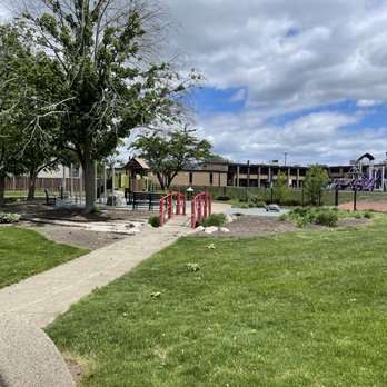 Overhill Park - A park with a grassy area, trees, and a concrete pathway leading to a small red bridge. There are benches near the trees and a playground with purple structures in the background, enclosed by a black fence. The sky is partly cloudy with patches of blue visible. - Playgrounds Near Me - Morton Grove, Illinois