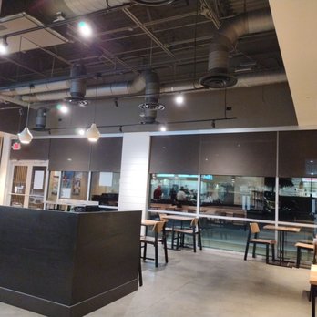 CAVA - Interior of a restaurant with wooden tables and chairs, large windows with closed blinds, and visible ceiling vents and lighting fixtures. - Mediterranean Near Me - Georgetown, Texas