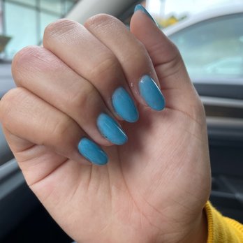 Red Nails - Cuticles not cleaned up. - The image shows a person's left hand with neatly painted blue nails, positioned inside a vehicle. The background includes a blurred view of parked cars and building windows, indicating the photo was taken in a car. The person is wearing a yellow sleeve, and the focus is on the well-manicured nails and the hand's natural skin tone. - Nail Salons Near Me - Eagan, Minnesota
