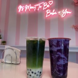 Green Thai Tea and Purple Sweet Potato Milk Tea with Tiger Walling - The image shows two bubble tea drinks on a light-colored table. The left drink has a green tea color with tapioca pearls at the bottom, while the right drink is purple with a marbled appearance. Behind the drinks, a pink-striped wall features a neon sign that reads 'G & M T F B Bubble - You' with a heart symbol, indicating a bubble tea shop setting.