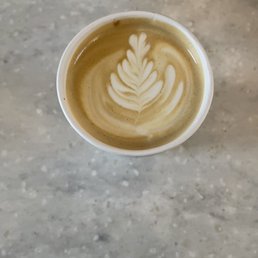Vanilla Latte - A top-down view of a latte in a white cup, featuring foam art with a leaf design. The latte is on a gray, textured surface, and the foam art is detailed with symmetrical leaf shapes.