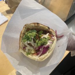 Falafel Pita Sandwich - A pita sandwich contains falafel, chopped vegetables, and green sauce, served in a paper wrapper.