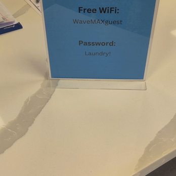 Wavemax Laundry - Aurora - Free Wi-Fi - A sign on a white table displays free Wi-Fi details for WaveMAX Laundry, including the network name 'WaveMAXguest' and the password 'Laundry!'. The sign is blue with wave graphics and is supported by a clear stand. The background shows part of a black chair, some blue laundry carts, and a small black box. - Laundromat Near Me - Aurora, Colorado