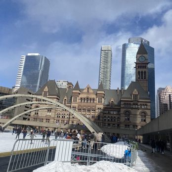 Nathan Phillips Square - City - The image shows Toronto's historic city hall with its unique curved design and clock tower, set against a backdrop of modern glass skyscrapers. The scene includes an outdoor ice skating rink with people skating and walking, some snow on the ground, and a partly cloudy sky overhead. The Canadian flag is visible on the right side of the image. - Landmarks & Historical Buildings Near Me - Toronto, Ontario