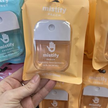 Five Below - A hand holds a yellow pouch containing a Mistify Peach Hydrating Hand Sanitizer, with a clear window showing the sanitizer bottle inside. The pouch is hanging on a display hook among other Mistify hand sanitizer pouches, which are in different colors. The product label indicates it is peach-scented and contains 1.2 fl oz (35 ml) of sanitizer. - Discount Store Near Me - Cedar Park, Texas