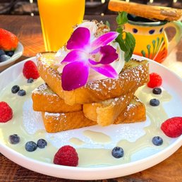 French Toast - French toast with whipped cream, edible flower, and powdered sugar, accompanied by berries and syrup on a wooden table.