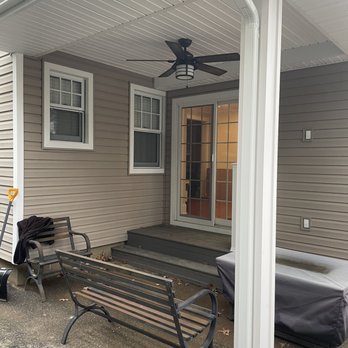 Murphy Construction - Side view off new family room - The image shows a covered outdoor patio area attached to a house with beige vinyl siding. The patio has a ceiling fan installed on the white ceiling. Two metal benches with wooden slats are positioned on the concrete ground, with some fallen leaves nearby. A sliding glass door provides access to the interior of the house, and there is a small set of wooden steps leading up to it. A covered outdoor furniture piece is partially visible on the right side of the image. - General Contractors Near Me - Oceanside, New York