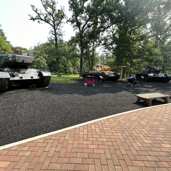 Cantigny - Three military tanks are positioned on a black gravel area in a park, surrounded by trees and benches, with a brick pathway in the foreground. - Museums Near Me - Wheaton, Illinois