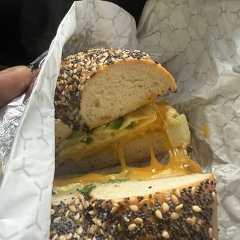 Bagel Boy - A bagel sandwich with cheese, lettuce, and other fillings, partially sliced, wrapped in branded paper. The bagel has a topping of sesame seeds and poppy seeds. The sandwich appears to contain melted cheese and fresh vegetables, with the bagel's interior visible. - Bagels Near Me - Brooklyn, New York