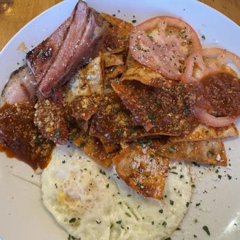 M'tucci's Italian - Chilaquiles - The image shows a white round plate with a serving of lasagna topped with tomato slices, alongside fried eggs garnished with herbs, and strips of bacon. The dish is presented on a wooden table, suggesting a casual dining environment. The food items are arranged with the lasagna in the center, the eggs at the bottom, and the tomato slices on the right side of the plate. - Italian Near Me - Albuquerque, New Mexico