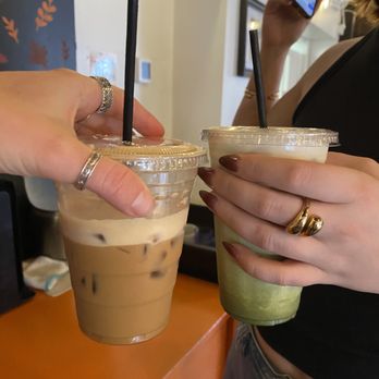 Haute Coffee - espresso (with oat milk) and matcha latte - Two people are holding iced coffee drinks in clear plastic cups with black straws inside a café. One drink appears to be a beige iced coffee with ice cubes, and the other is a green matcha latte. The person on the left has rings on their fingers, and the person on the right has rings and painted nails. In the background, a woman is using her phone, and the café interior features a wall with decorative elements and framed pictures. - Coffee & Tea Near Me - Concord, Massachusetts